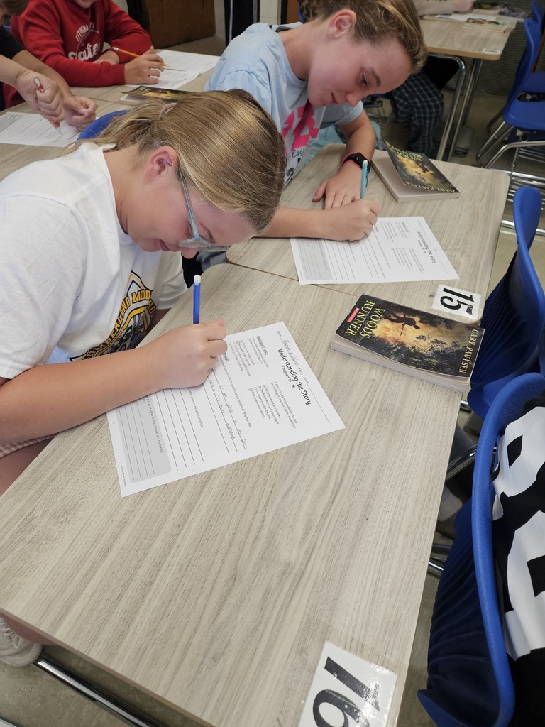 Two students are stiiting together at desks reading a book and filling out a paper.