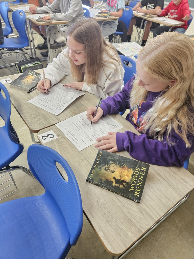 Two students are stiiting together at desks reading a book and filling out a paper.