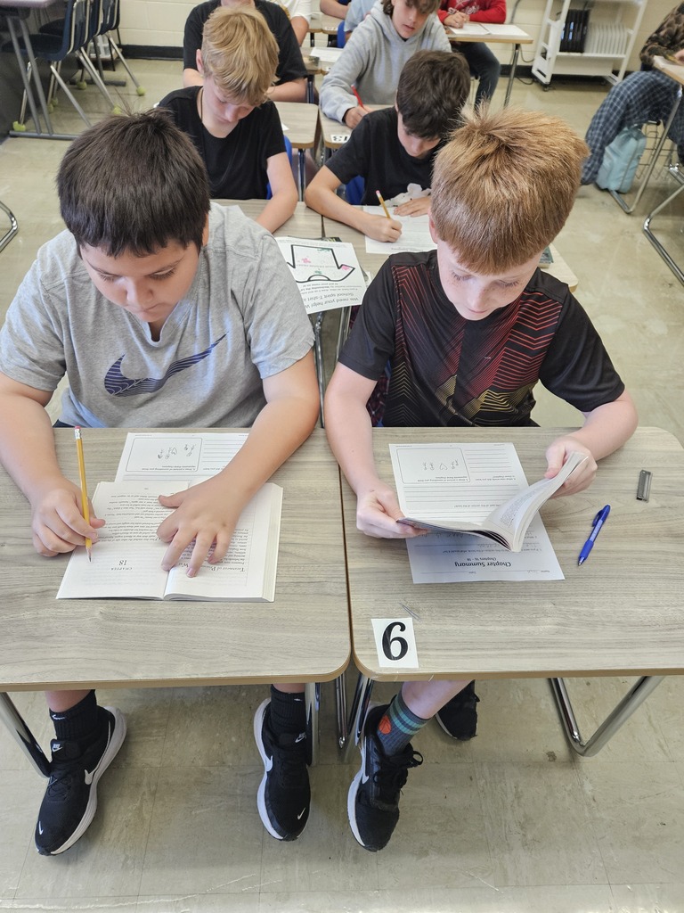 Two students are stiiting together at desks reading a book and filling out a paper.