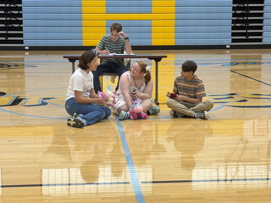 Four students are doing a skit. Three are talking sitting on the ground. The fourth is doing work at a table. 