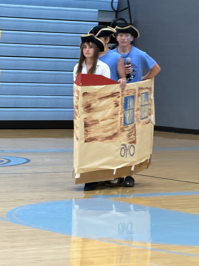 Three students are doing a skit. They in a cardboard ship walking out to center court. 