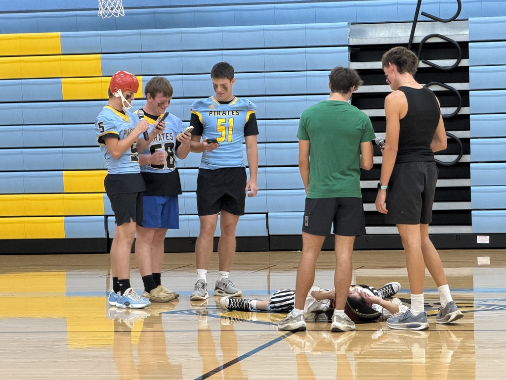 Five students are doing a skit. One is lying on the floor with two looking on. Three are wearing football uniforms and reading lines.