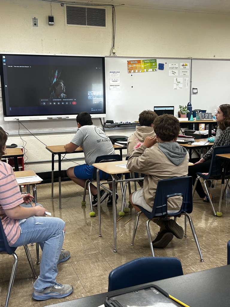 Students are in desks facing the SmartBoard. A person is on a Google meet and talking to the students.