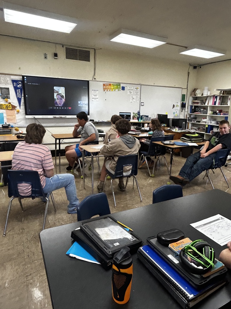 Students are in desks facing the SmartBoard. A person is on a Google meet and talking to the students.