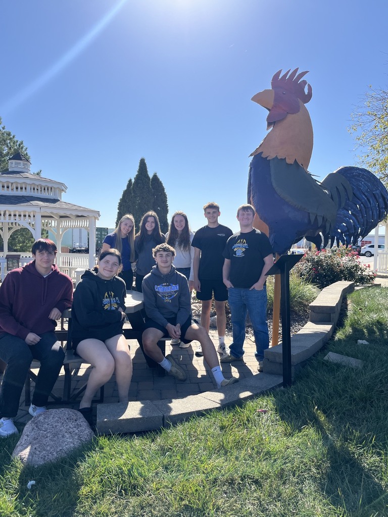 Eight students are pictured - three sitting and five standing. There is a large rooser and gazebo on the background.