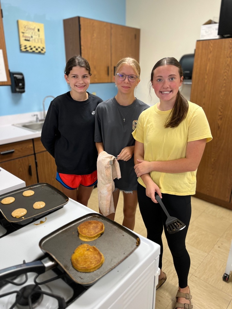 Three students are standing in front of an oven. One has a spatula and an other has a towel. There are two pans.