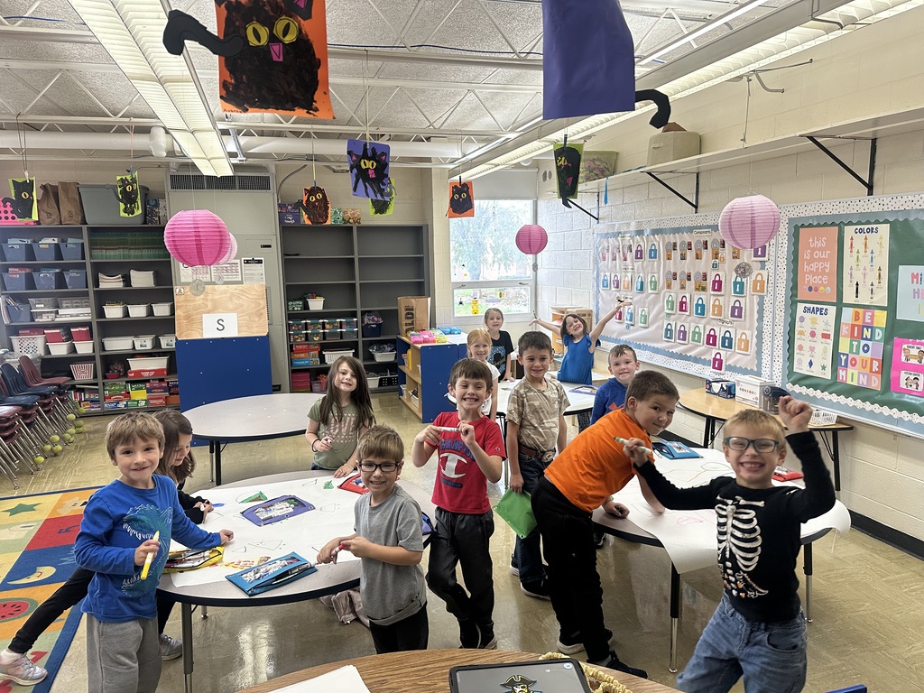 The class is in groups of four or five at three round tables. They have markers and are drawing on a large butcher block paper.