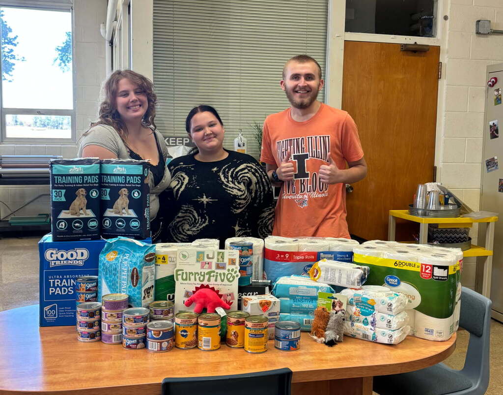 Three students are standing behing a table full of pet products like, food, wipes, paper towels, and toys.
