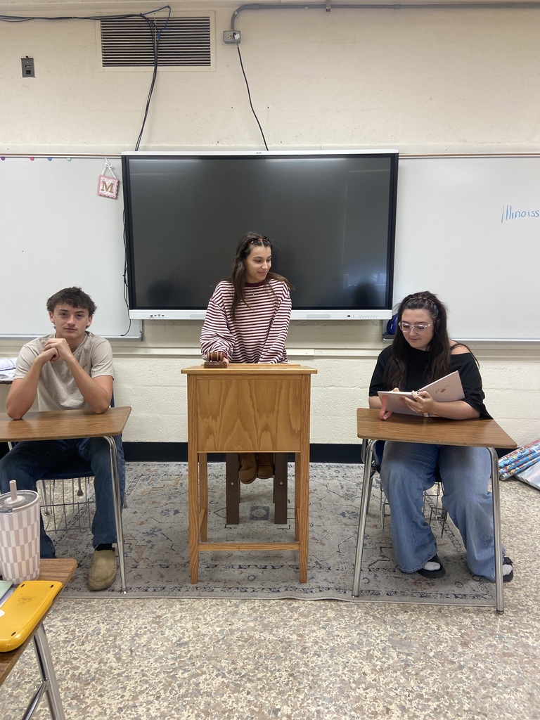 Three students are featured. Two are acting as attorneys and are sitting at desks. One is the judge and is staning behind a podium. 