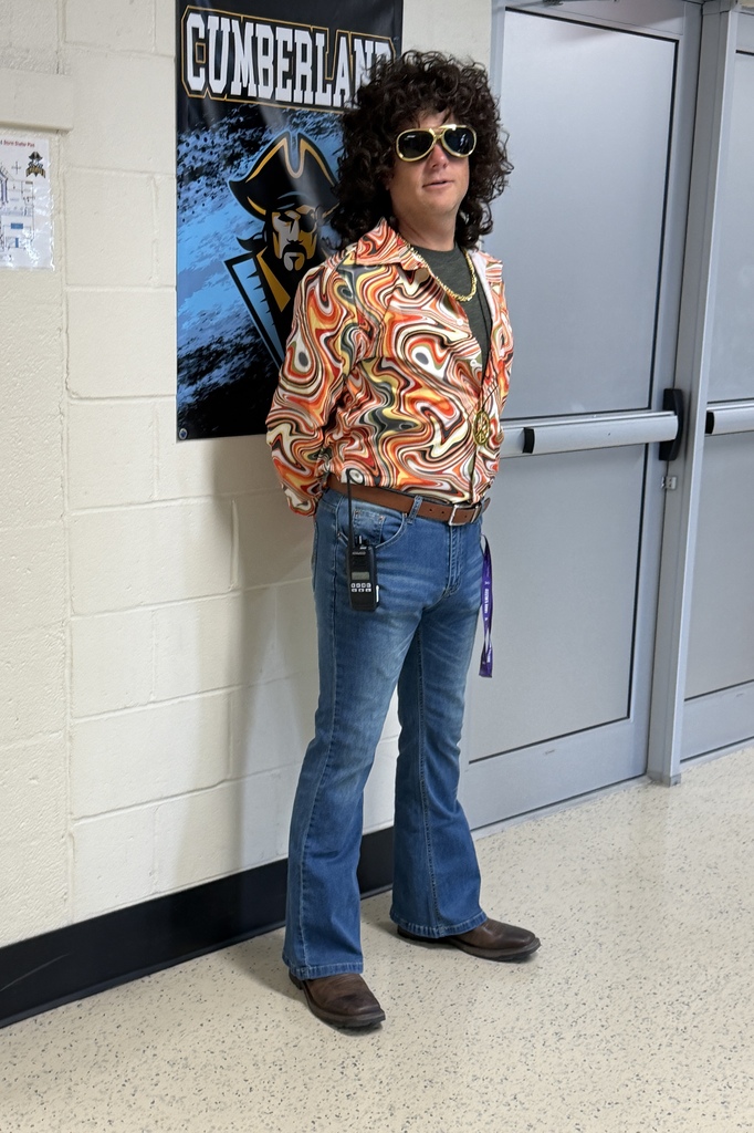A principal is dressed from the 70s with jeans, a wig, and a tye dye shirt. 