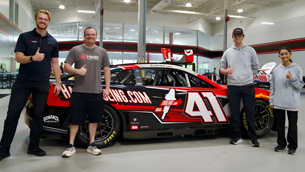 A group of four people standing in a garage next to a black and red NASCAR race car with the number 41 and HaasCNC.com branding. The car is positioned indoors with bright overhead lighting and visible workshop equipment in the background.