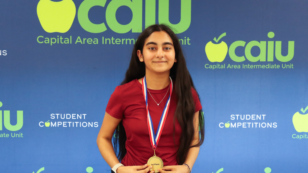 Eagle View sixth grader Ishika Talreja poses with a first place medal at the PA Media and Design Regionals with a CAIU background behind her.