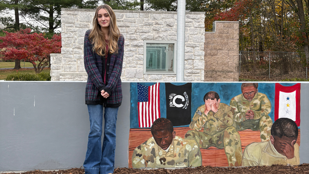 CVHS junior Alexis Mayers-Davies stands beside a mural she created for the Veterans Memorial Wall Art Project, featuring soldiers and patriotic flags.