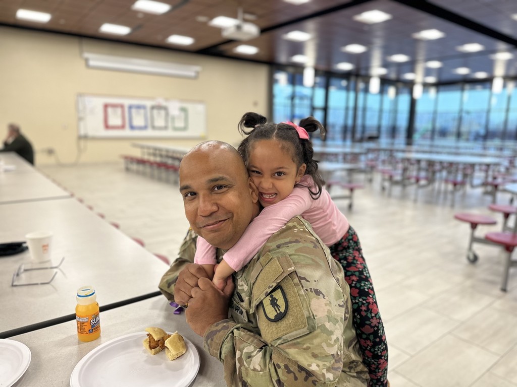 Little girl hanging on her dad's back, eating breakfast at Winding Creek Elementary to celebrate Month of the Military Child.