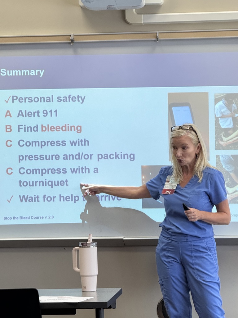 Woman in blue scrubs pointing at a slide on bleeding emergency steps.