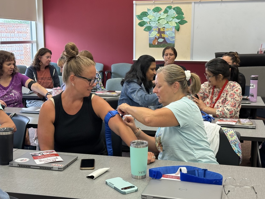 Person applies a blue tourniquet to another's arm in a classroom.