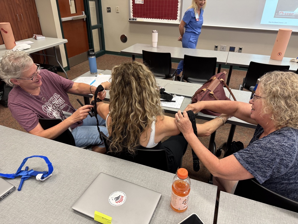 Three people practice applying a tourniquet in a classroom with an instructor observing.
