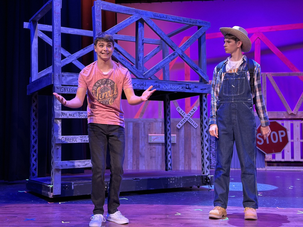 Two young performers on stage during a dress rehearsal for Footloose: Youth Edition. The set features a rustic, rural design with wooden structures and warning signs. One performer wears a pink t-shirt and black pants, gesturing expressively, while the other is dressed in overalls, a plaid shirt, and a straw hat. The scene is lit with purple and blue theatrical lighting.