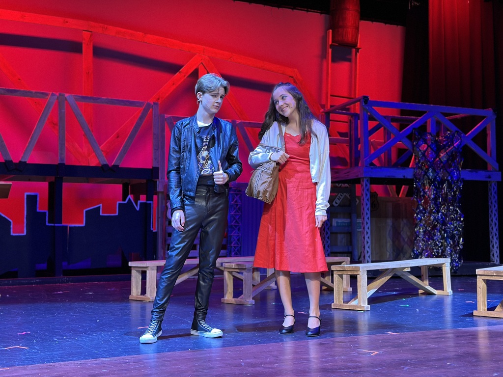 Two young actors on stage during a dress rehearsal for Footloose: Youth Edition. One wears a black leather jacket, black pants, and sneakers, while the other wears a red dress with a white cardigan. The set includes wooden benches and a red-lit backdrop evoking an industrial or urban atmosphere.