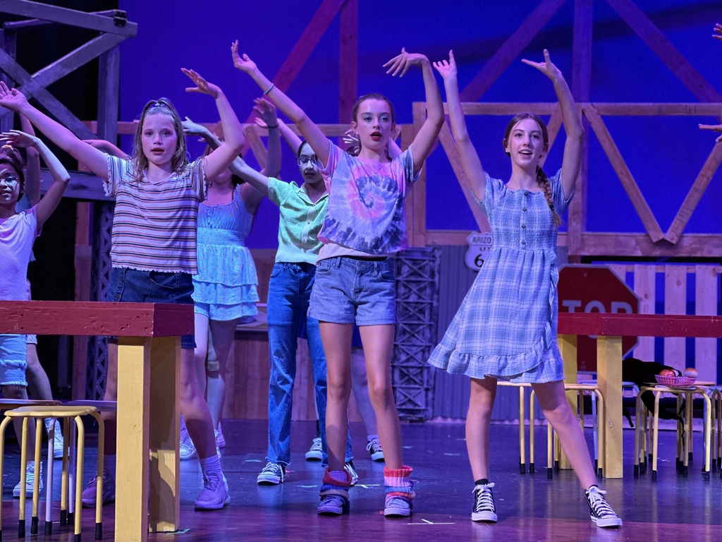 A group of young performers dancing on stage during a dress rehearsal for Footloose: Youth Edition. They stand in a line with arms raised and spread out, wearing casual outfits like t-shirts, shorts, and dresses. The set includes wooden structures and a stop sign, with bright lighting highlighting their energetic choreography.