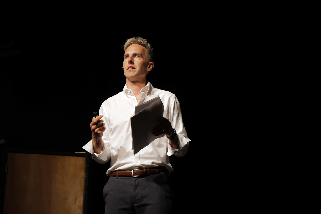Person in white shirt speaking on stage to a crowd, holding papers.