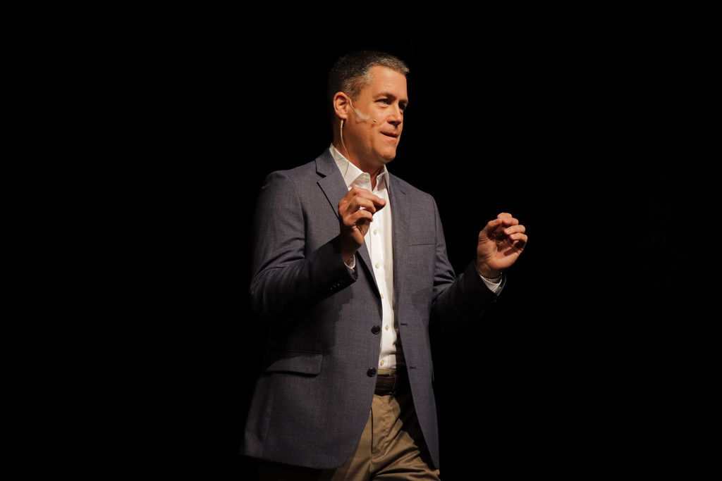Person in grey blazer speaking on stage to a crowd, gesturing with both hands against a dark background.