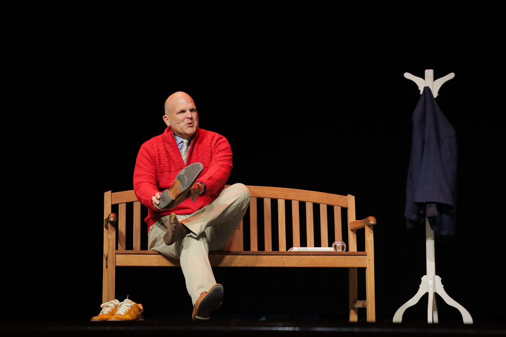 Person in red sweater sitting on a bench putting on shoes, with yellow shoes nearby and a coat rack in the background.