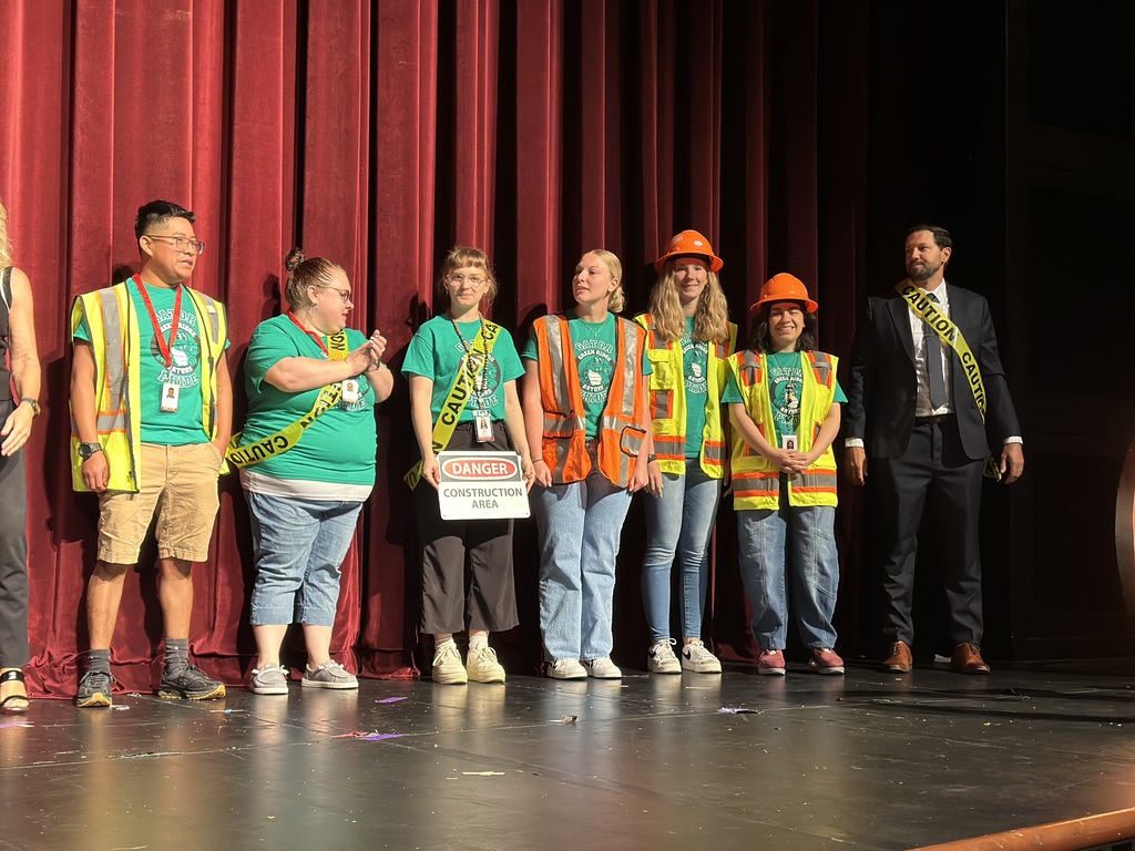 Group on stage in front of red curtains, wearing construction gear and holding a 'Danger Construction Area' sign