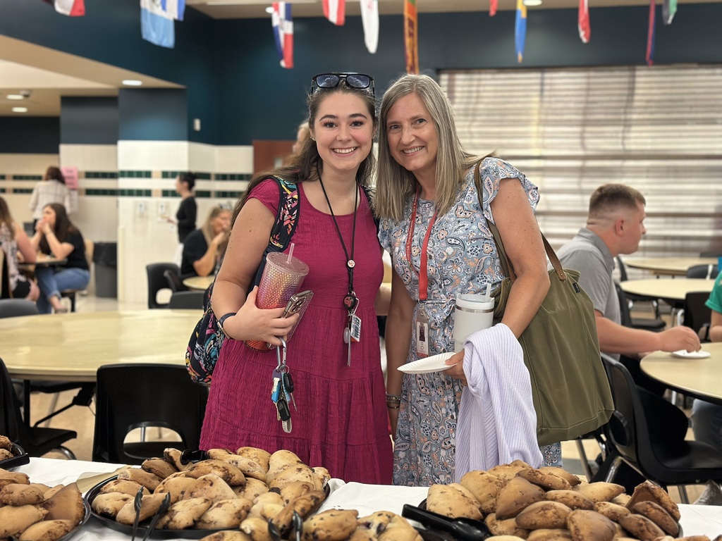 Two people standing by a pastry table in a cafeteria, with international flags hanging above.