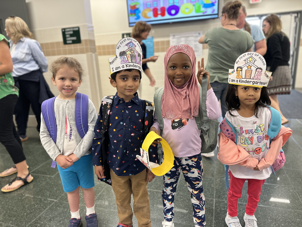 Four children with backpacks holding hands in a school hallway, two wearing first-day crowns.