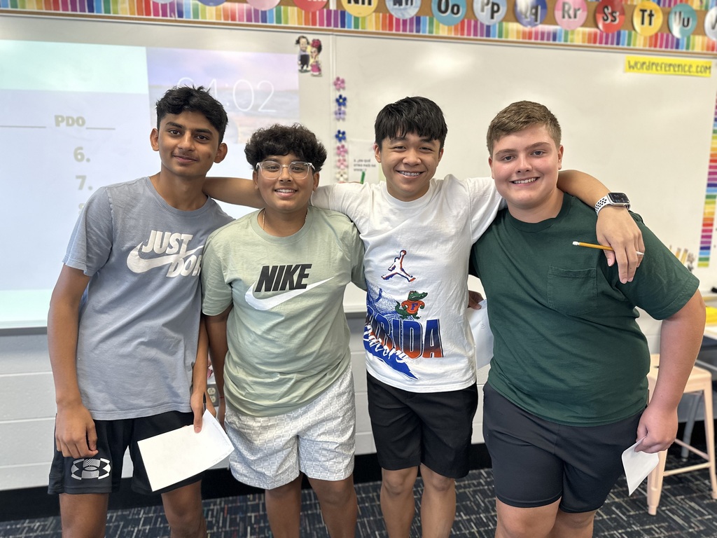 Four students standing together in a classroom with papers and alphabet decorations in the background.