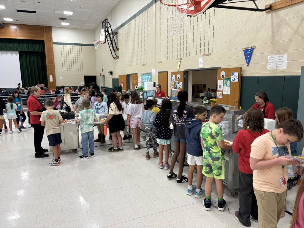 Children stand in line at a school cafeteria inside a gymnasium with basketball hoops and a stage in the background.