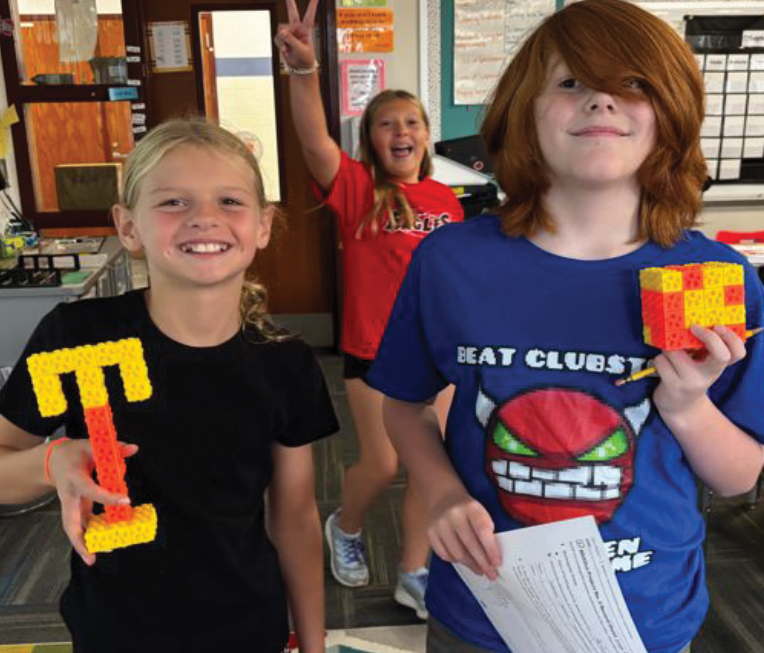 Students display 3D-creations, including a letter 'T' and a cube, with a classmate flashing a peace sign in the background
