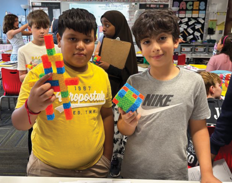 Two students display colorful block creations in a classroom filled with desks and learning materials.