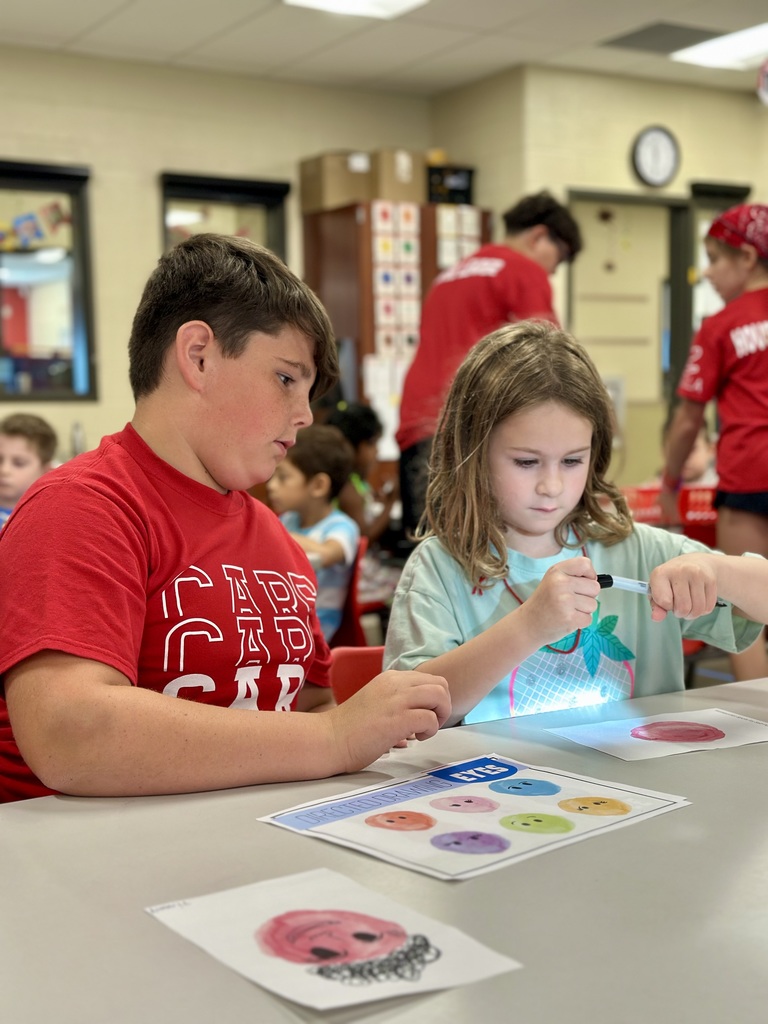 Two children seated at a classroom table working with colorful circular images, with others and educational materials in the background.