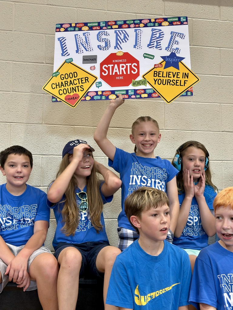 Children in blue 'INSPIRE' shirts sit together, with one holding a sign promoting kindness, character, and self-belief.