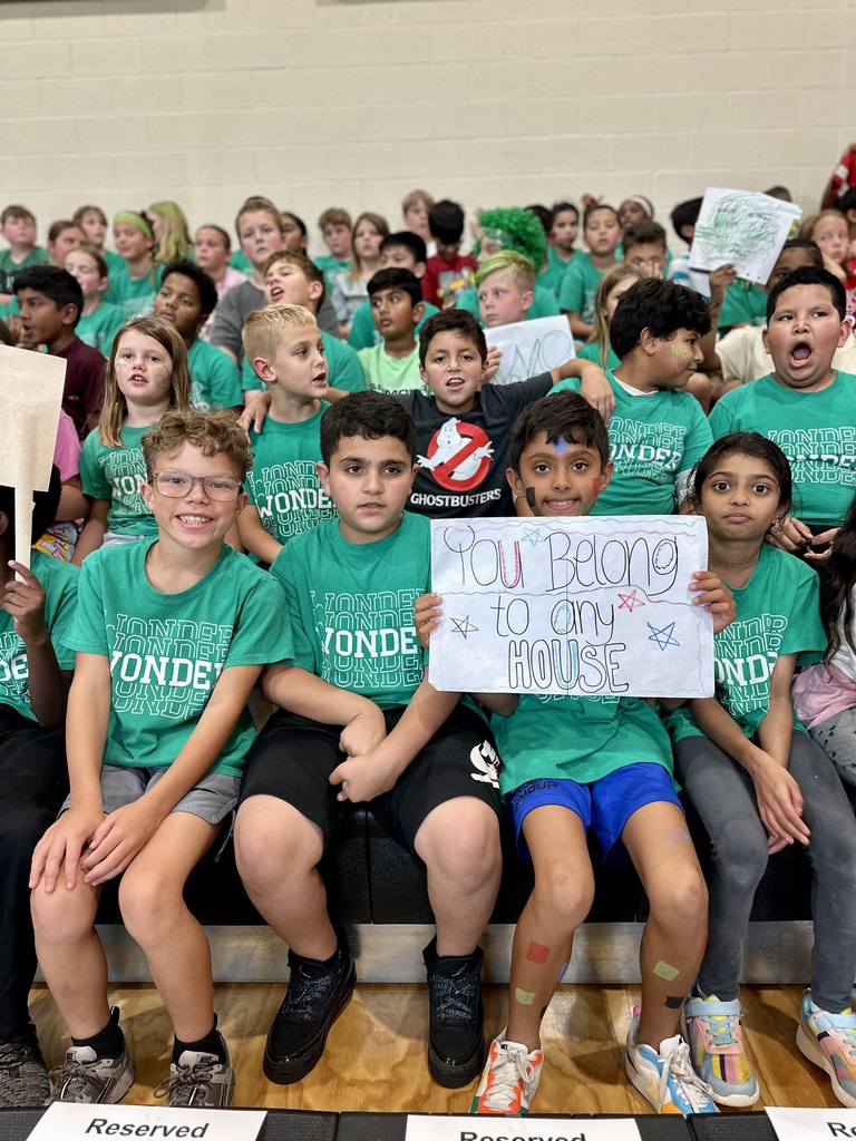 Children in green 'WONDER' shirts sit in a gym, with one holding a sign that says 'You belong to any HOUSE.'