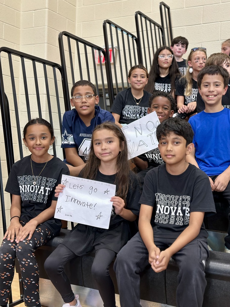 Children in 'INNOVATE' shirts sit on bleachers, holding supportive signs for their school house.