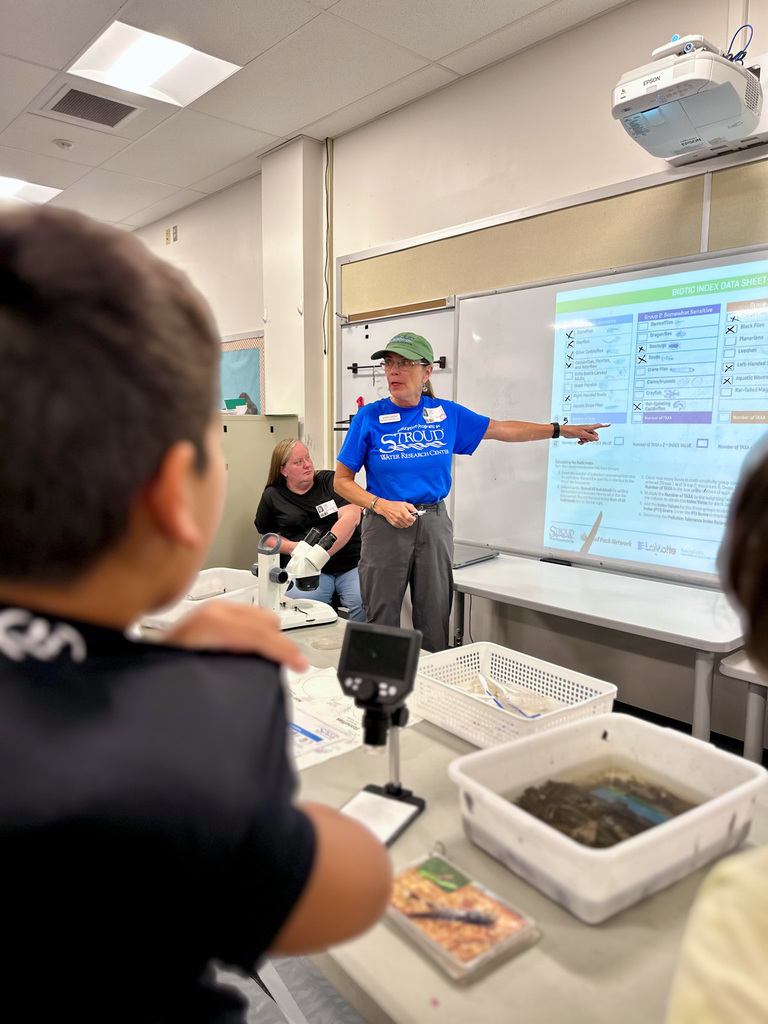 Stroud Water Research employee pointing at a projected screen while students work with microscopes and materials in a classroom.