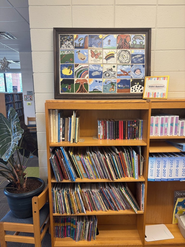 A wooden bookshelf in a school library filled with children's books. On top sits a framed collage of colorful student artwork, including rainbows and butterflies. A sign reading "Biddle's Bookshelf" is displayed next to the artwork. Additional labeled shelves and a potted plant on a chair are visible nearby.