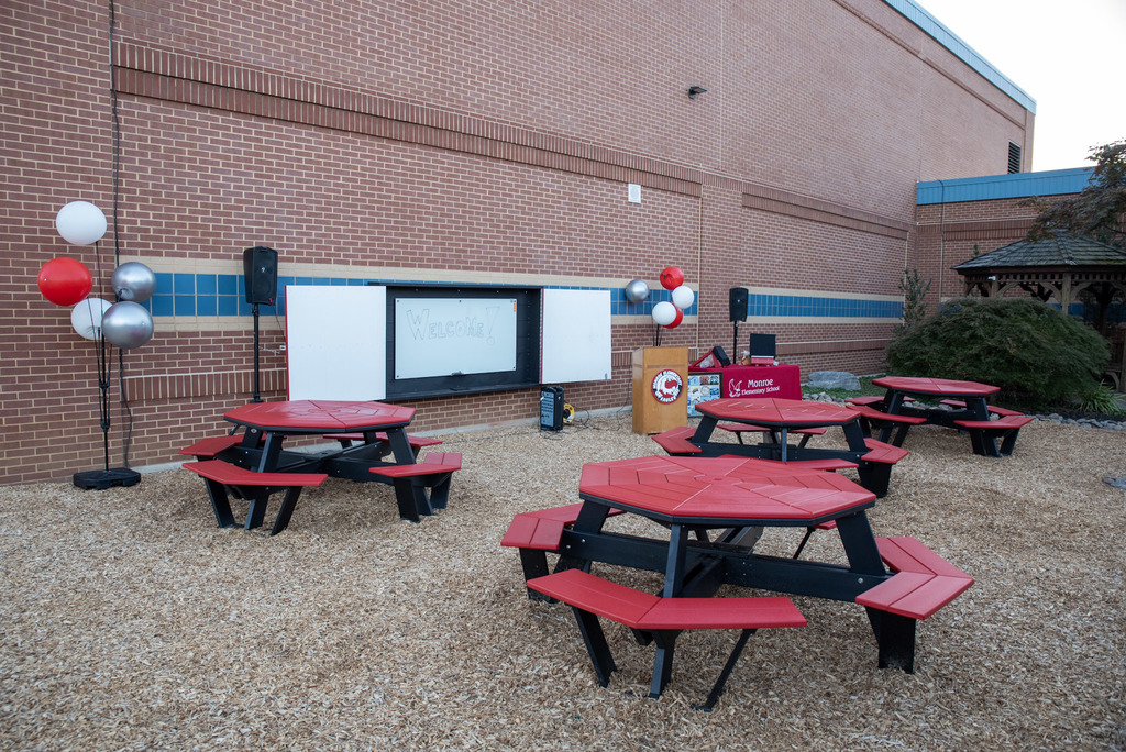 An outdoor classroom area featuring red picnic tables on a bed of wood chips, surrounded by a garden bed with rocks and plants. A paved walkway runs through the space, with a large tree and greenery in the background. A brick school building with a blue and yellow stripe and an information board is visible to the right.