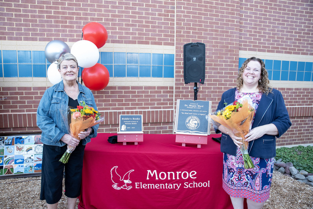 Two individuals stand smiling in front of a red tablecloth that reads "Monroe Elementary School" with an eagle logo. Each holds a bouquet of flowers. Behind them is a brick wall with blue tile accents and a cluster of red, white, and silver balloons. On the table are two plaques labeled "Biddle's Bookshelf" and "Dr. Megan Ward Outdoor Classroom," along with a speaker. The scene is part of an outdoor dedication event.
