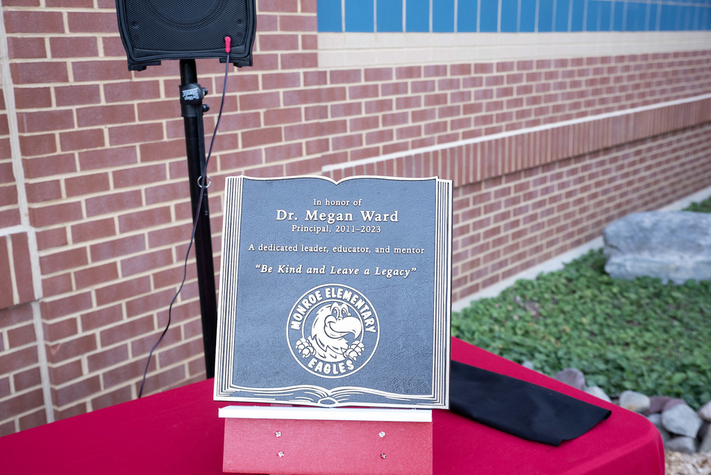 Plaque honoring Dr. Megan Ward, Principal from 2011–2023, with the message 'Be Kind and Leave a Legacy' and the Monroe Elementary Eagles emblem. Displayed on a red tablecloth with a brick wall and greenery in the background.