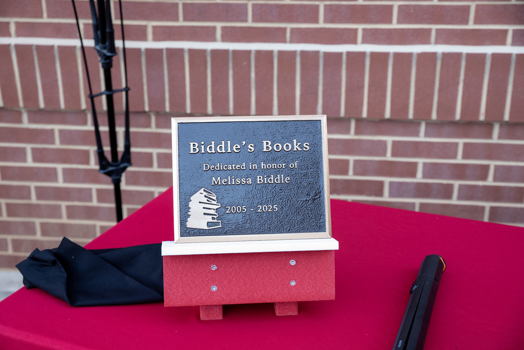 Plaque titled 'Biddle's Books' dedicated in honor of Melissa Biddle, 2005–2025, featuring an illustration of a stack of books. Displayed on a red tablecloth with a brick wall background.