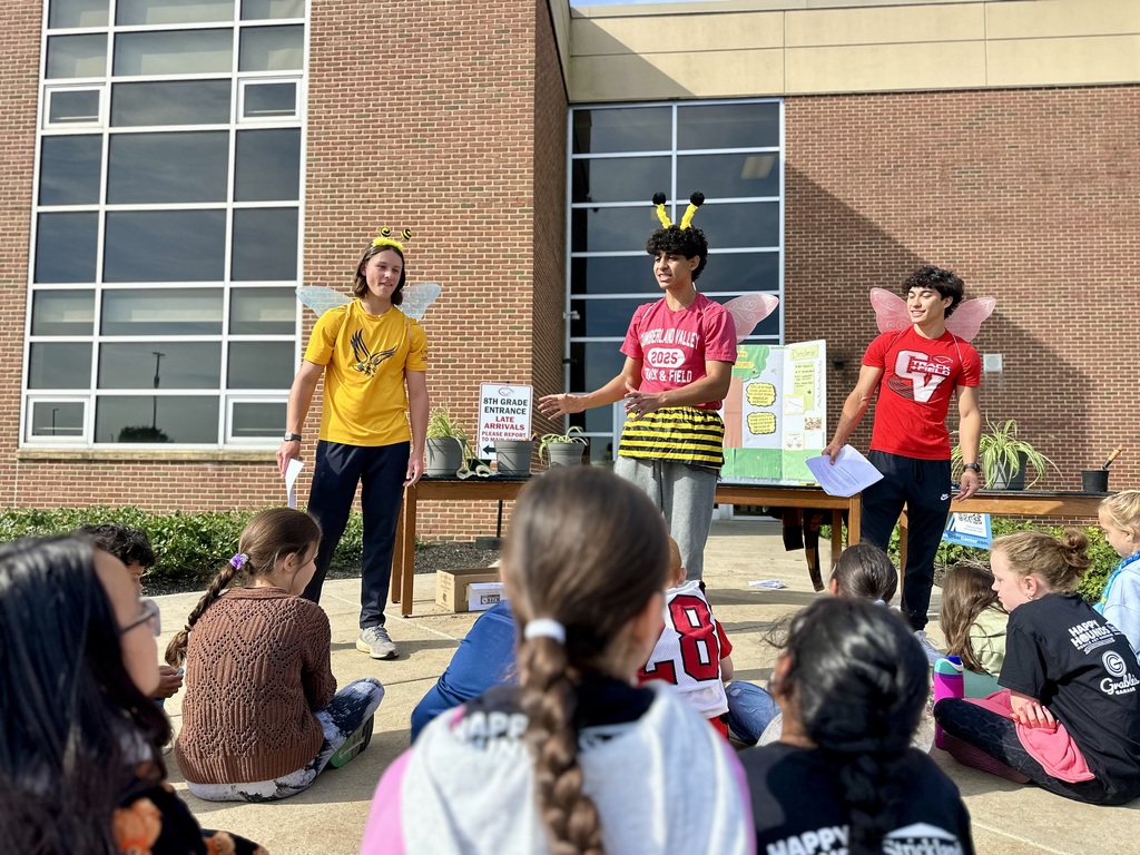 Students sit outside a building listening to three high school presenters dressed as bees and butterflies, with a table of plants and nature posters behind them.