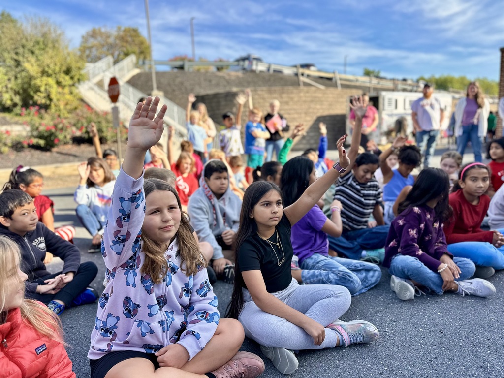 Children sit on the ground outdoors, some raising their hands, during an activity near a stone wall with greenery and observers in the background.