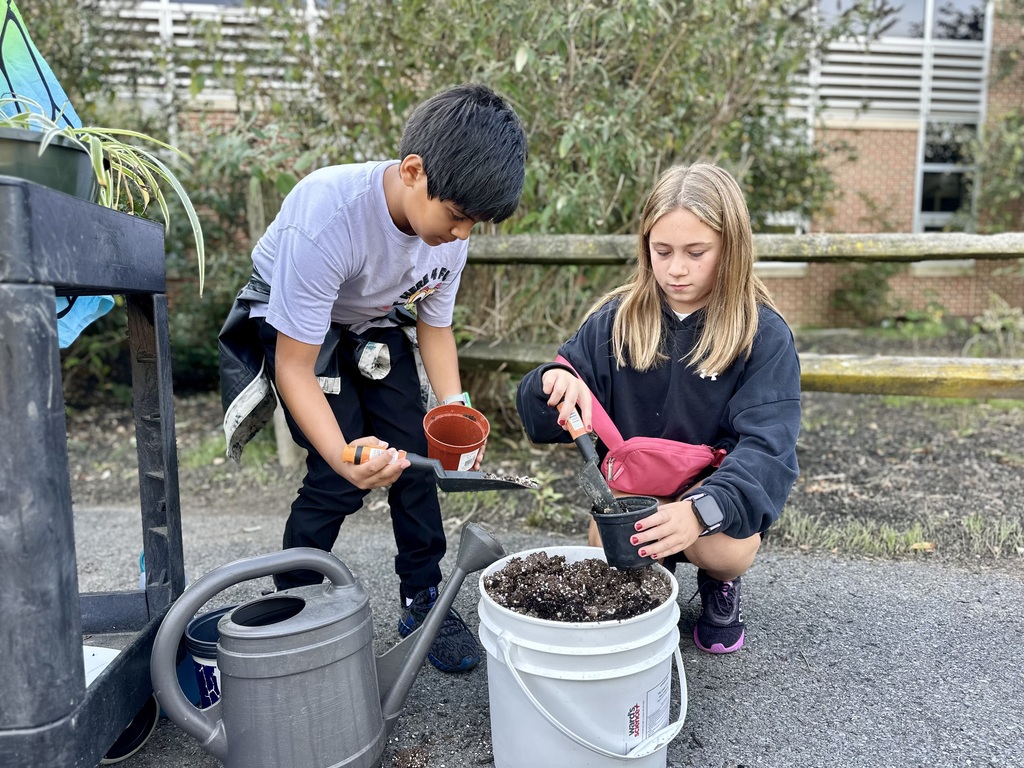 Two children garden outdoors, using a scoop and trowel to fill pots with soil near a watering can and cart of plants.