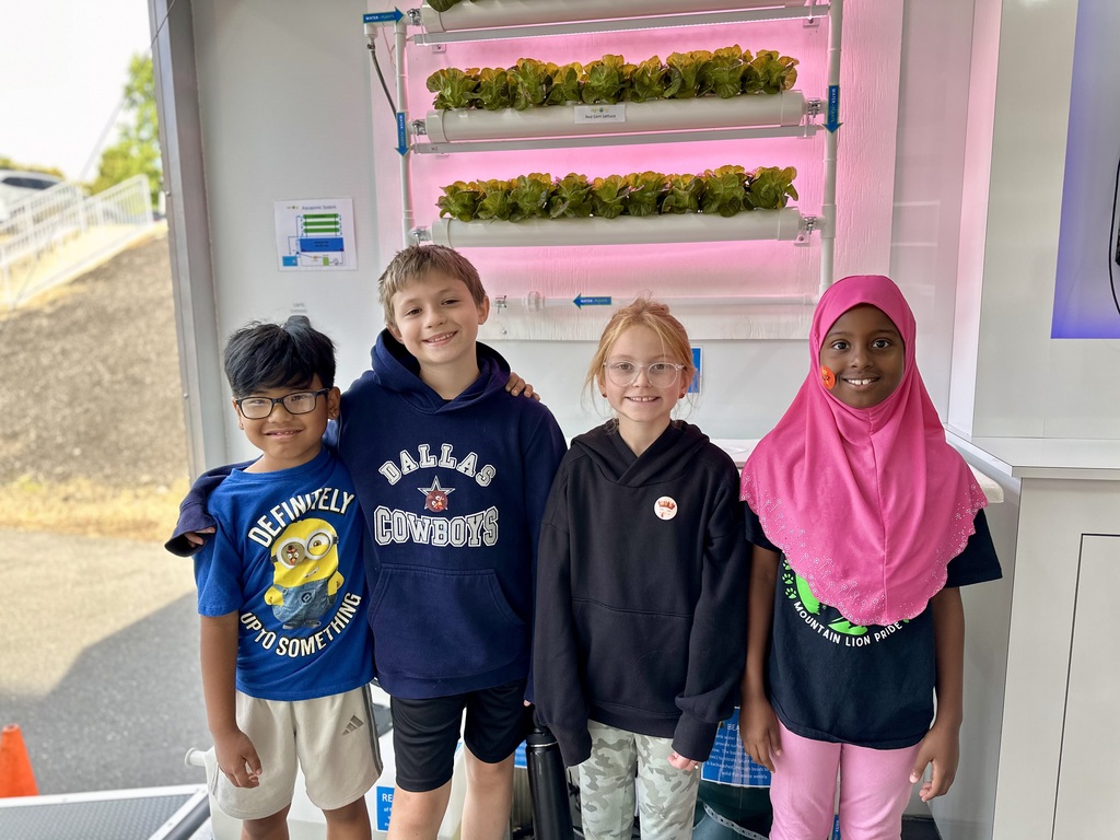 Four children stand in front of a vertical hydroponic garden with leafy greens under pink LED lights