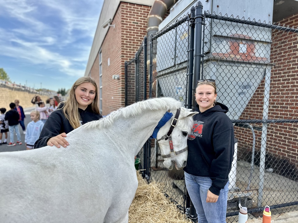 Two high school students stand beside a white horse wearing a blue halter on straw bedding.