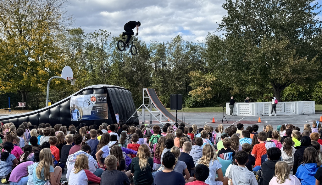 Monroe Elementary students sit outdoors watching a BMX stunt show. A BMX rider soars high in the air above a ramp while students look on in excitement. The show takes place under a partly cloudy sky with trees showing early fall colors in the background.